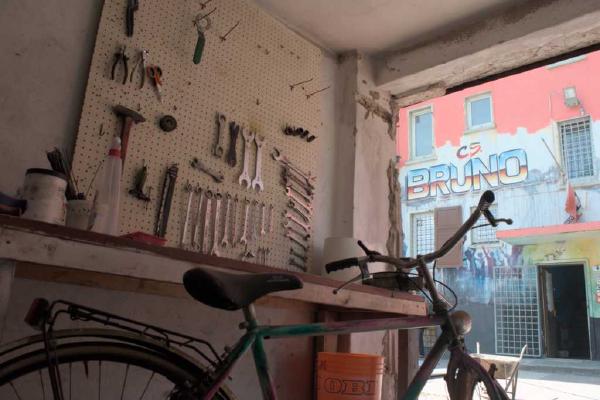 Una foto scattata dall'interno di Ciclostile, la ciclofficina popolare del Centro Sociale Bruno di Trento. In primo piano si vede una bicicletta appoggiata al con gli attrezzi. Sullo sfondo si vede l'ingresso del centro sociale con il murale 'CS BRUNO'.